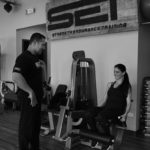 black and white image of male trainer with female client on leg extension machine